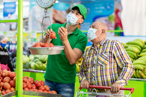 Latin man of average age of 30 years old employee of a grocery store attends to elderly man who is a customer of the store who asks for help to choose the best foods, both wear masks to avoid contagion of covid-19