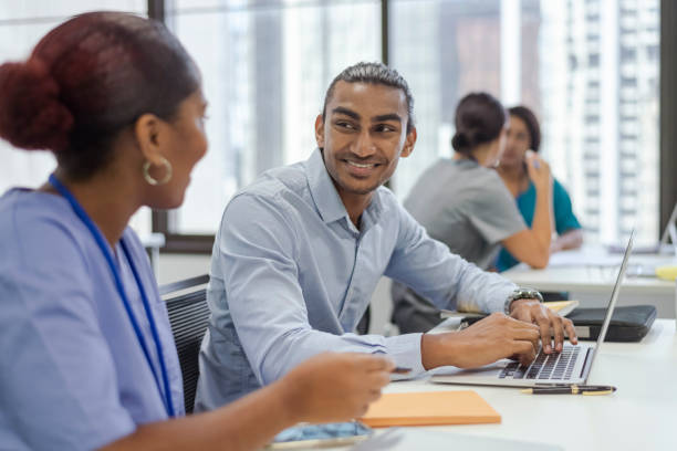 Medical students work together on a research project Shot focused on a medical student of Indian descent smiling at colleague. Multi-ethnic group of medical students in a modern classroom setting. All students sitting at a desk. Using laptop. Wearing medical scrubs. university students australia stock pictures, royalty-free photos & images