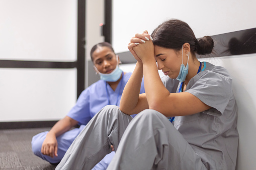 A distressed female nurse is sitting in a hospital hallway with her head in her hands. She is stressed and exhausted from the overload of patients in the ICU unit. A female colleague sits next to her to comfort her.