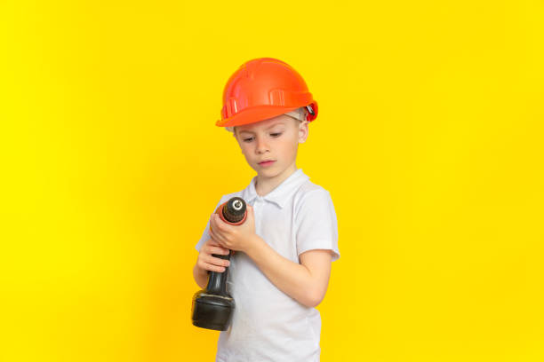 istock Building craft training. A boy in a kaskuke stands on a yellow background and looks attentively at a construction electric tool. The profession is a builder, the development of children. 1320407247