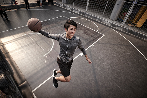 young asian adult male basketball player attempting a slam dunk on outdoor court