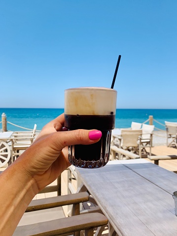 Glass with iced coffee with milk in female hand in beach cafe