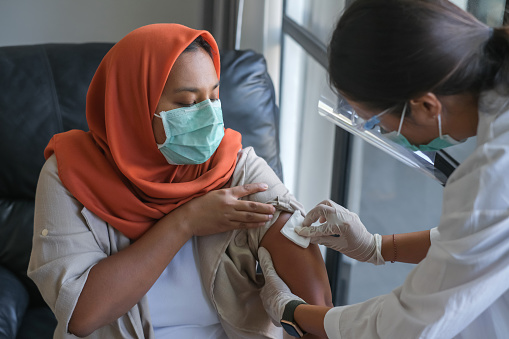 Close-up shot of Indonesian healthcare worker rubbing an alcohol wipe on the patient’s upper arm. The patient is wearing an orange hijab and surgical mask.