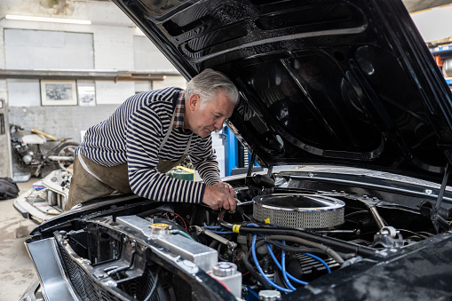 Waist-up view of Caucasian man wearing apron over casual clothing and leaning in to make adjustments to classic car engine.