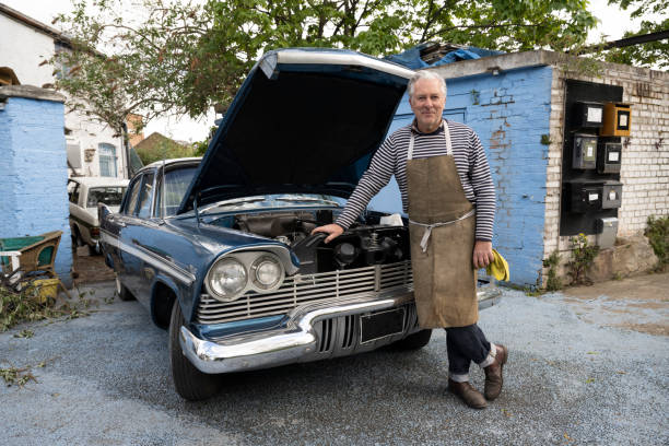 Proud restorer of classic 1950s American sedan Outdoor full length portrait of senior Caucasian man in work apron smiling at camera while standing in front of car with hood up. vintage car stock pictures, royalty-free photos & images