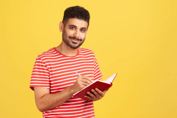 istock Happy positive man with beard in striped t-shirt making notes with pen at notepad, writing down information and looking at camera with smile 1320463668