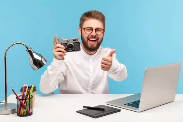 Cheerful excited cameraman showing thumbs up holding vintage photocamera and smiling looking at camera, sitting at workplace, satisfied with photo shoot. Indoor studio shot isolated on blue background