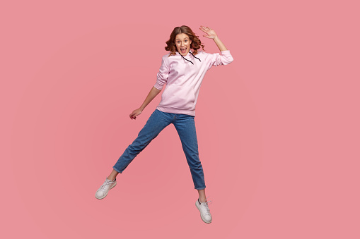 Full length portrait of cheerful young female in hoodie and jeans with toothy smile waving hand in jump, feeling happy, greeting. Indoor studio shot isolated on pink background