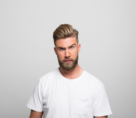Portrait of handsome bearded young man wearing white t-shirt, staring at camera. Studio shot, grey background.