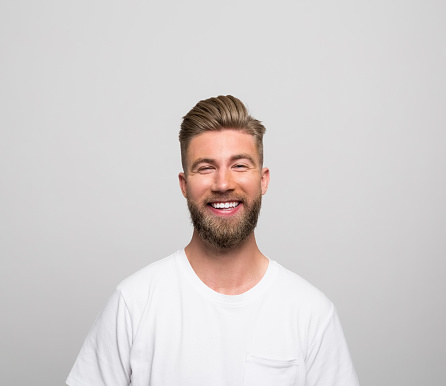 Portrait of handsome bearded young man wearing white t-shirt, smiling at camera. Studio shot, grey background.