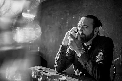 Young handsome Caucasian man with beard drinking a coffee in cafeteria and talking on phone. Black and white.