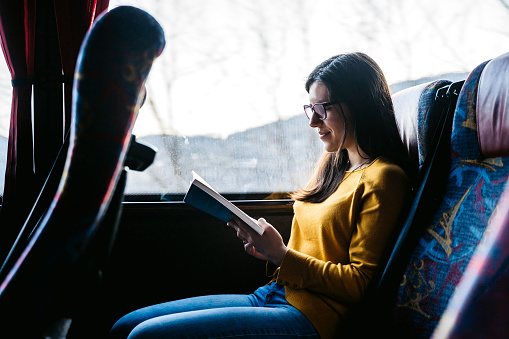 Young beautiful Caucasian woman reading book in bus on a road trip.