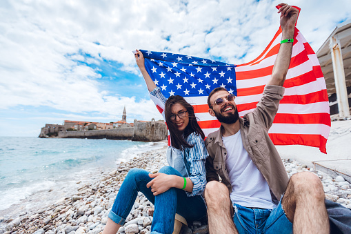 Young caucasian  couple holding American flag and enjoying at nice day on beach.