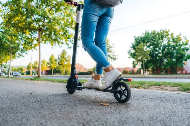 Close up on woman legs feet standing on the electric kick scooter on the pavement wearing jeans and sneakers in summer day. Back view riding.
