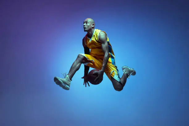 In motion. Portrait of athletic african-american male basketball player training isolated in neon light on blue background. Concept of health, professional sport, hobby. Passionate, fashionable. Dynamic