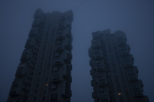 Two tall residential buildings in the form of a tower, immersed in a misty blue sky. Cyberpunk stylistics. Dramatic, depressing mood.