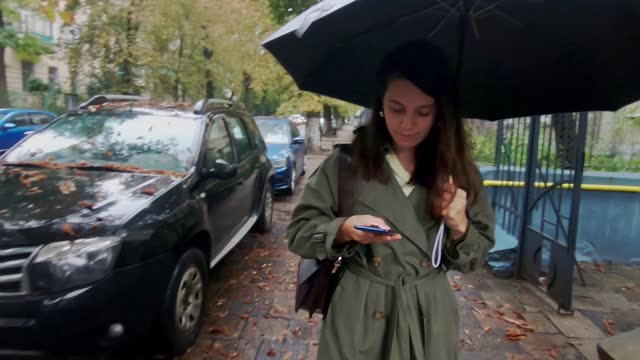 young pretty woman walking with phone by rainy street under umbrella
