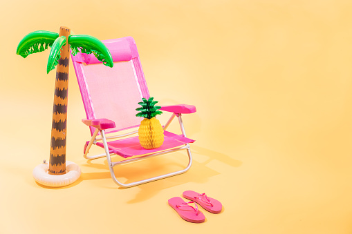 Pink beach chair with a paper pineapple on top of it, pink flip-flops and an inflatable palm tree on yellow background. 3D illustration