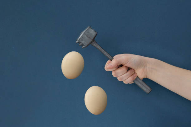 istock Female hand holds iron hammer ready to hit light brown eggs flying on dark blue background 1320488522