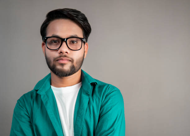 Portrait of a bespectacled Indian young man looking at the camera. Portrait of a bespectacled Indian young man standing against the isolated gray background and looking at the camera with a blank expression. blank expression photos stock pictures, royalty-free photos & images