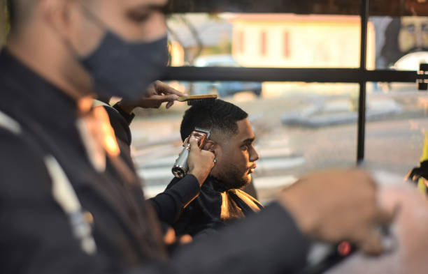 Young man having a haircut at a barbershop Portrait of a young black Brazilian man sitting at a barbershop in São Paulo city, Brazil, having a haircut. The barbers are wearing a social uniform and face mask, due to Covid-19 protection measures, as they do their work. Before the young black man there is another customer and another barber working, defocussed. Behind the young black customer there is a glass wall, with a view to the street, defocussed. black barber stock pictures, royalty-free photos & images