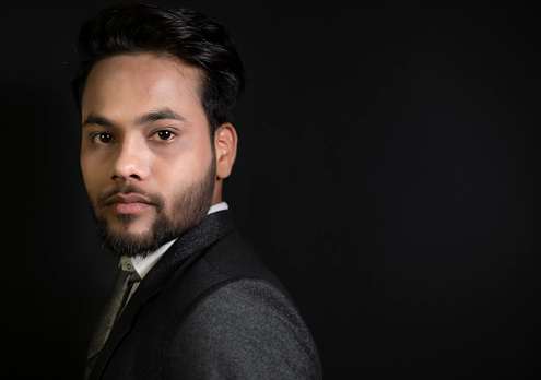 Low key side view portrait of handsome, elegant, fashionable Indian young businessman informal standing against a dark black background and looking at the camera with a blank expression.