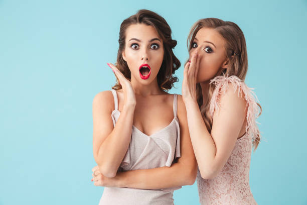 фотография двух сплетни девочек 20-х носить платья и партии макияж говорить секреты в ухо и выражая удивление, изолированные на синем фоне - beautiful pink blue lifestyles стоковые фото и изображения