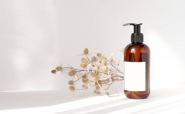 istock Brown bottle with dispenser and empty white label on light background. Next to shampoo package is bouquet of dried flowers. Rays of sunlight fall into background, leave shadows from objects 1320496541