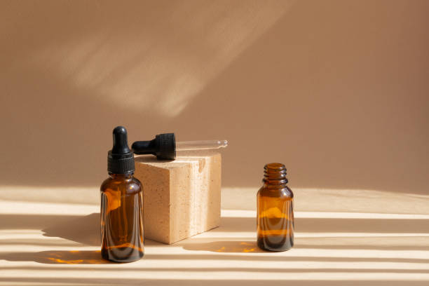 istock Two glass bubbles on brown background, in rays of sunlight, cast shadow on the surface. One bottle with black cap, cap-pipette from second, lies on square stand. Concept of the beauty industry 1320496548
