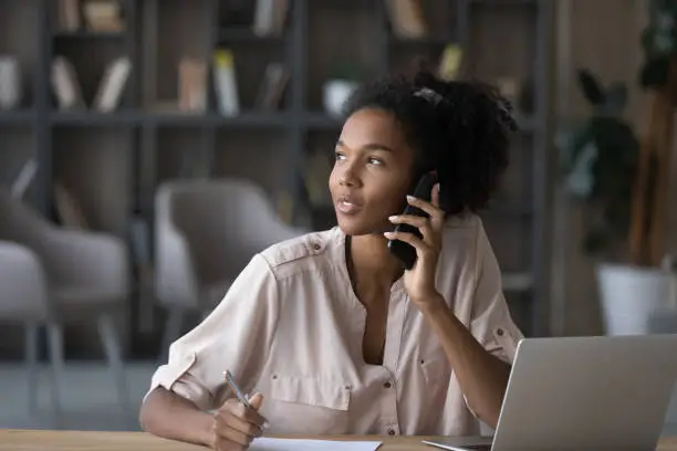 Dreamy young african american woman looking in distance, holding telephone conversation, giving professional consultation to client working in modern home office, distant communication concept.