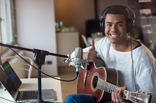 Afro american young man male musician sitting and smiling on chair practicing learning playing guitar using his laptop, professional music equipment with microphone and headphone at home. photo