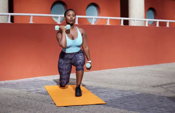 Photo of Shot of a young woman on a gym mat using dummbells against an urban background