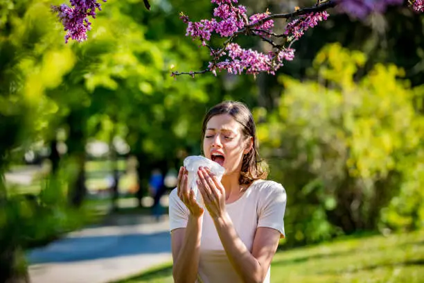 Photo of Young pretty woman blowing nose in front of blooming tree. Spring allergy concept