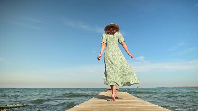 A young woman in a green summer dress and a straw hat in slow motion walks along a wooden pier in the morning on the sea during the wind. Walk grace beauty and femininity
