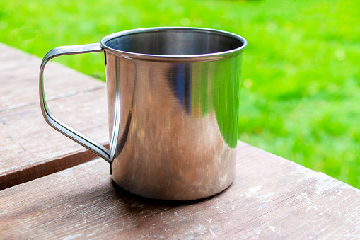 Metal mug with tea or coffee on wood table. Close-up of travel cup in camping with copy space.
