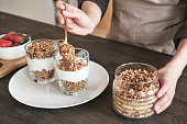 Close-up of unrecognizable woman standing at kitchen counter and putting muesli into mug while preparing granola for breakfast