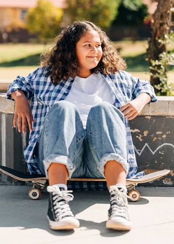 Young girl with a skateboard sitting at the skatepark. Female in casuals sitting on her skateboard looking away and smiling taking a rest from skating