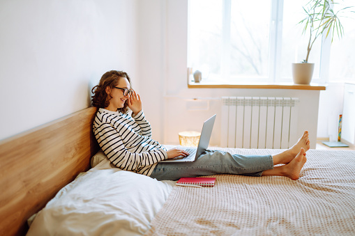 Education and working online. Young female freelancer sitting at comfortable bed in cosy home interior and working remotely on modern laptop computer. Online shopping in web store.