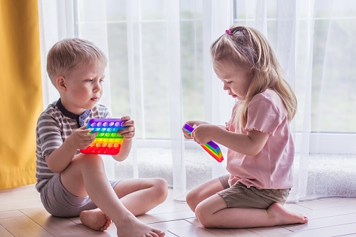 Blonde boy and girl children sit on the floor near the window and play silicone toy antistress pop it. Pop it sensory toy. Stress relief. Colorful anti stress silicone sensors toy