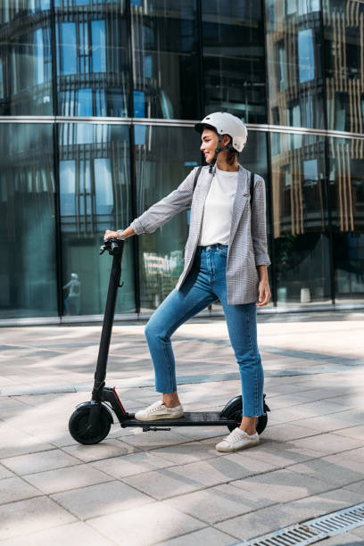 Side view young woman standing on electrical scooter near an office building. Female in helmet ready for a ride on scooter. Side view young woman standing on electrical scooter near an office building. Female in helmet ready for a ride on scooter. push scooter stock pictures, royalty-free photos & images