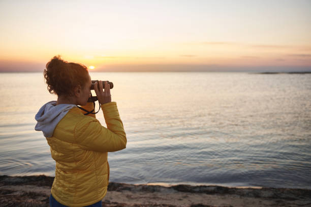 istock Young woman in a yellow jacket looking through binoculars and enjoying a beautiful view of the coastal landscape 1320541966