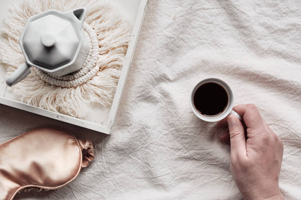 istock Espresso in hand, tray with white ceramic espresso coffee maker on trendy macrame pad. Sleep mask on crumpled bed sheets. Female hand with cup of espresso. Good morning Simple minimal top view 1320542119