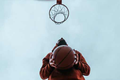 Male basketball player standing under basketball hoop. He is getting ready to score and practicing at basketball court in cloudy day