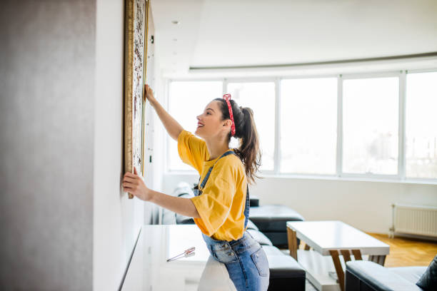 mujer joven colgando un cuadro en una pared con una mirada de concentración - decorar fotografías e imágenes de stock