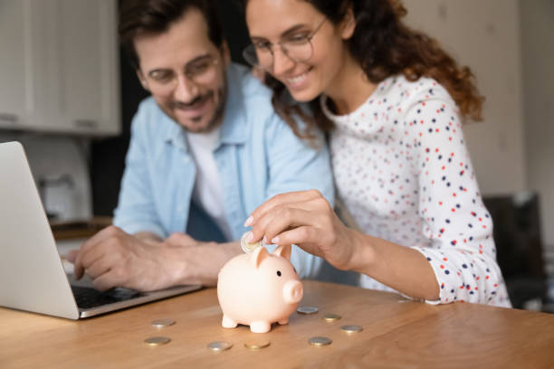 Happy millennial family couple planning future investment. Happy millennial family couple putting coins in piggybank, planning vacation or investments together, saving money for life insurance, managing future expenditures together using computer apps. financial wellbeing stock pictures, royalty-free photos & images