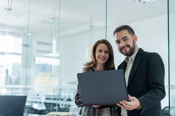 istock coworkers are standing inside the office working looking towards the camera while holding a laptop in their hand 1320567728