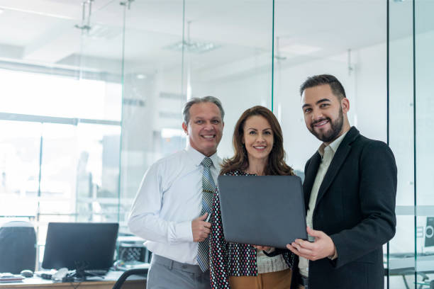 istock coworkers are standing inside the office working looking towards the camera while holding a laptop in their hand 1320567771