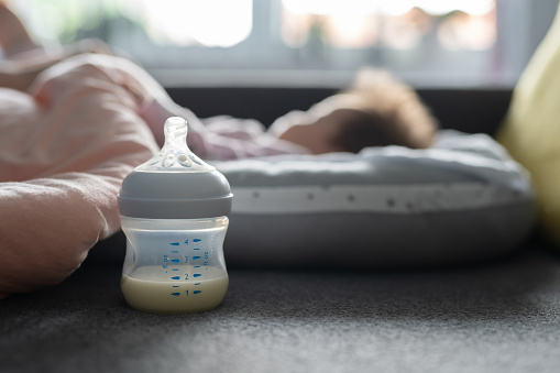 Close up selective focus on breast or formula milk in the baby bottle on the bed at home with sleeping baby infant in background - newborn feeding and nursing concept concept copy space
