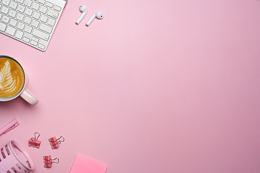 Feminine workspace with coffee cup, keyboard, wireless earphone and copy space on pink background.