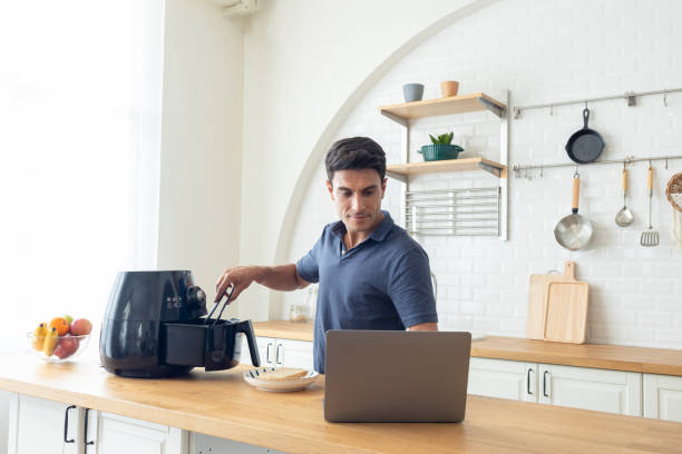 A portrait of a Hispanic man  learning the air fryer machine manual on the Internet in home. Adult men looking for recipes on laptop at modern kitchen.  He learning to cook with online course stock photo